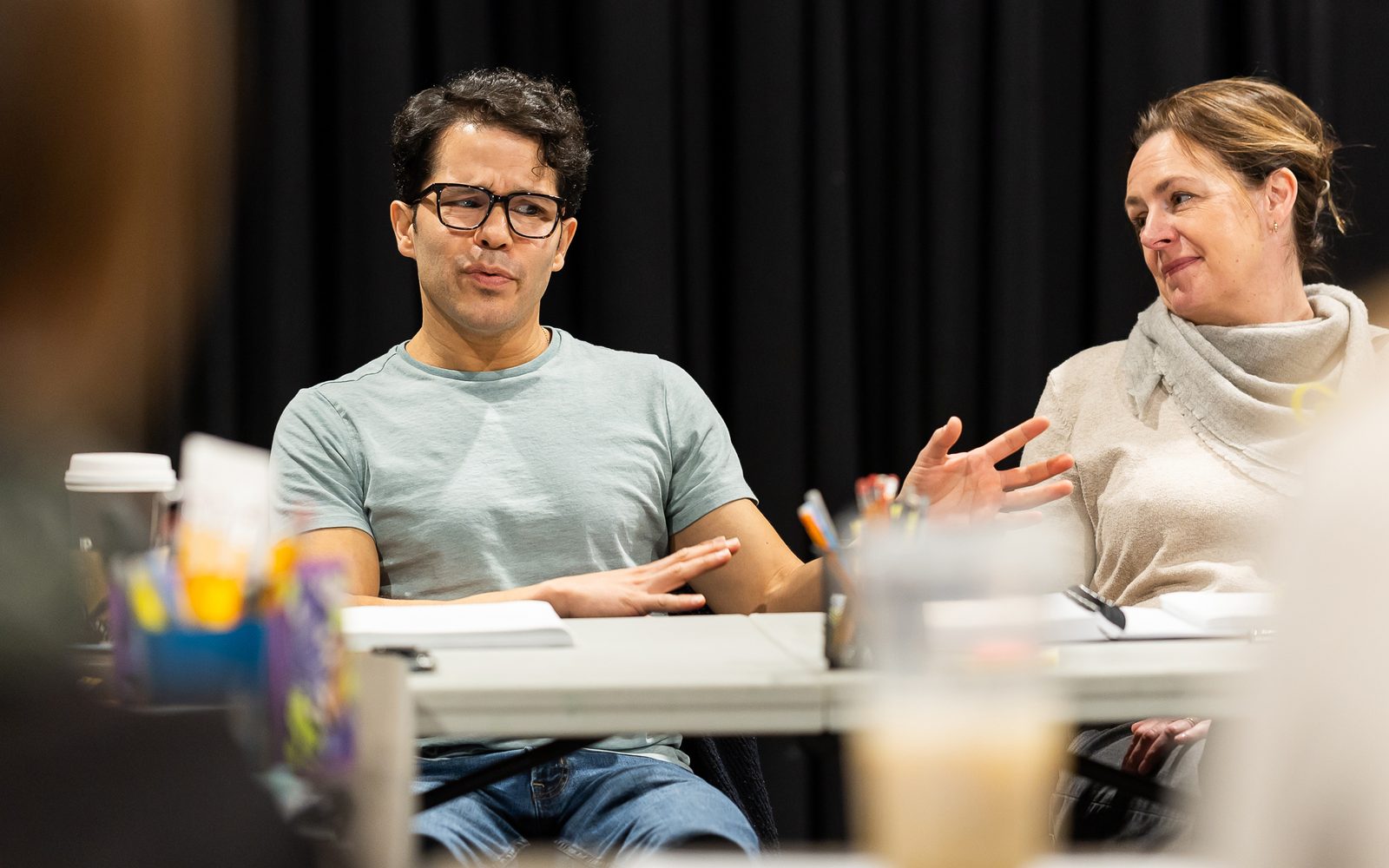 Carlo Albán in black glasses and a teal T-shirt speaks while sitting at a table, while Andrus Nichols in beige on the right watches him.
