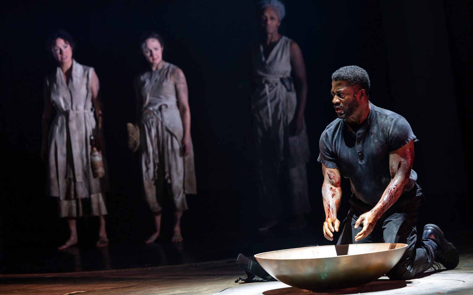Wayne T. Carr kneels before a gold bowl of water to wash blood off his hands. Alejandra Escalante, Kate Hamill, and Nike Imoru, dressed in white, dirtied robes, stand behind him and watch.
