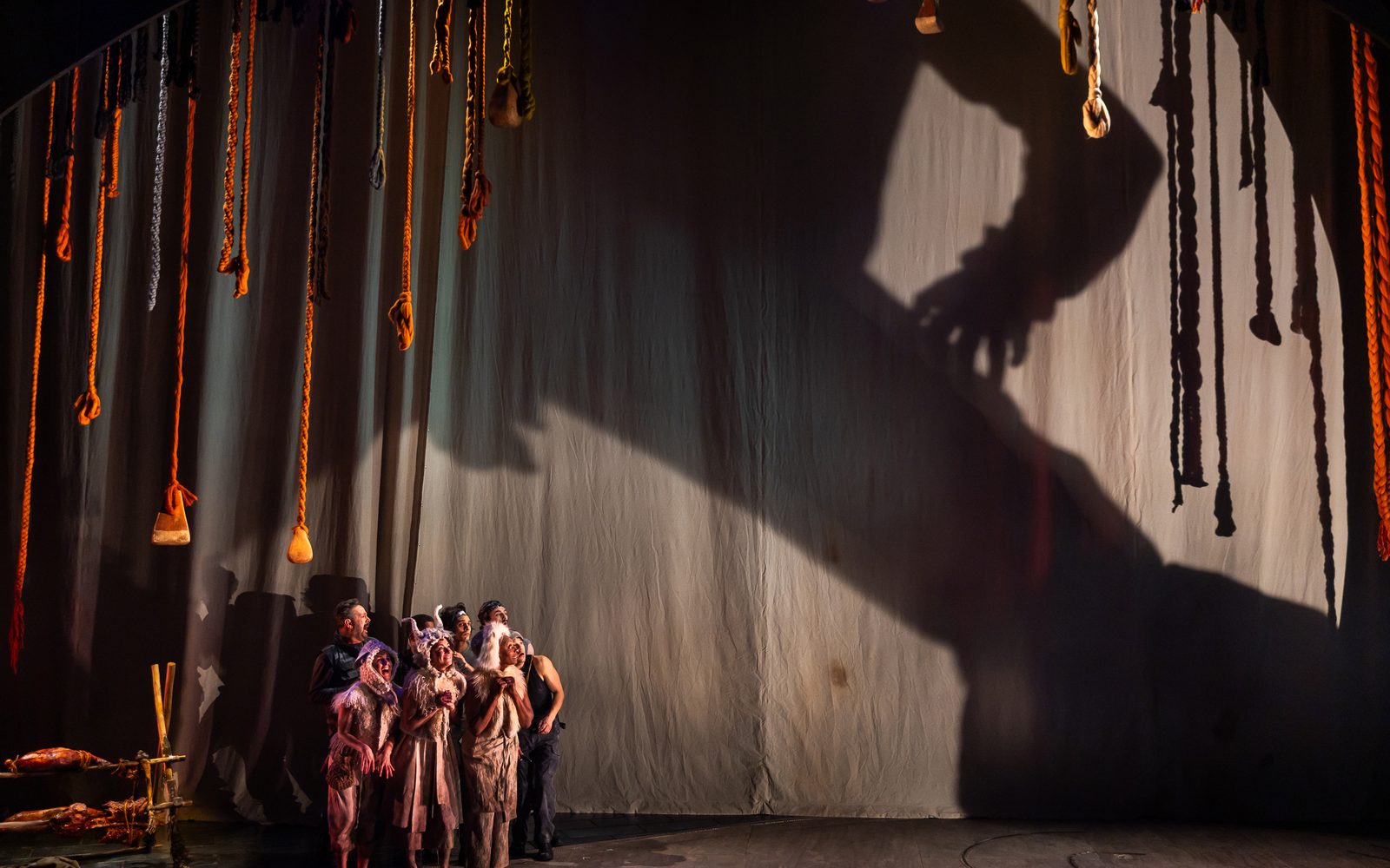 A shadow of a giant stands over the actors, who are grouped to one side underneath its legs.