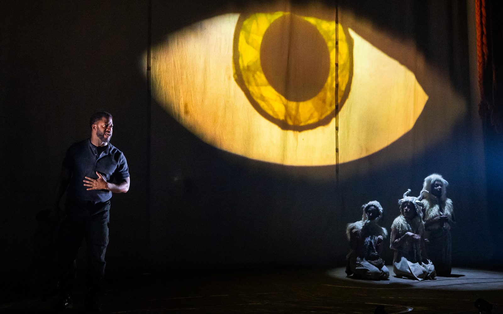 Wayne T. Carr stands to one side of a projection of a giant yellow eyeball, which watches over Kate Hamill, Alejandra Escalante, and Nike Imoru who are dressed as sheep.