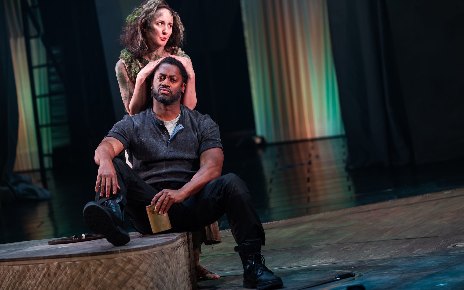 Kate Hamill in moss-covered blouse and dirt on her skin stands behind Wayne T. Carr with her hands on his head. He sits on a low platform in a dirty, dark blue henley shirt holding a flask.