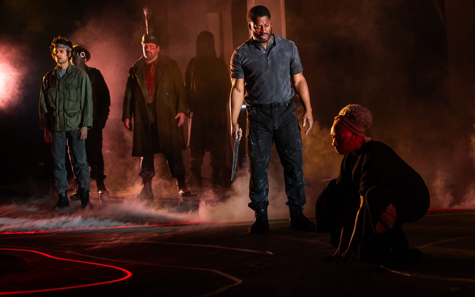 Keshav Moodliar, Chris Thorn, and other masked actors stand in red-tinged fog behind Wayne T. Carr, who holds a long dagger by his side and stares at Nike Imoru crouched on the ground.