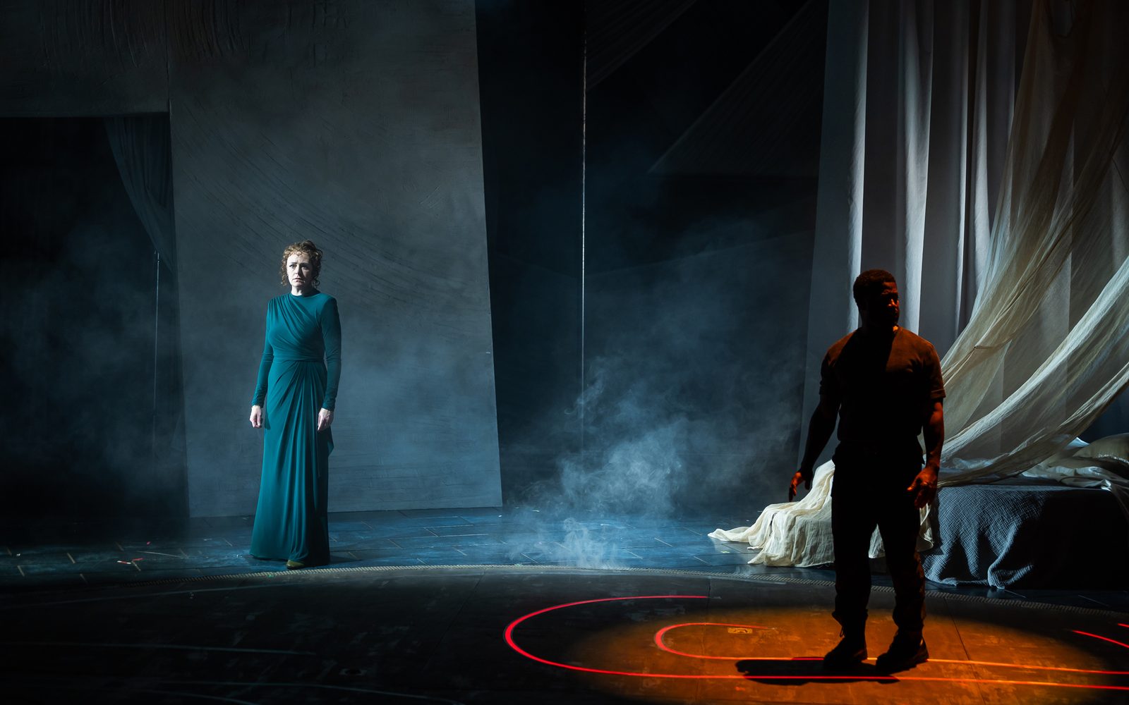 Andrus Nichols in a dark teal gown stands to the left with a worried expression on her face, while Wayne T. Carr in dark clothing stands on the right looking over his shoulder under a dim red light.