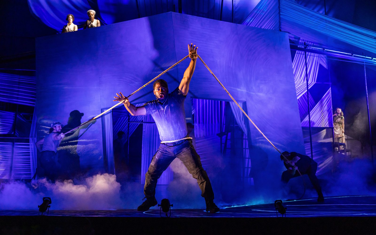 Wayne T. Carr stands center stage lit in a blue light holding a rope above his head as Benjamin Bonenfant and Jason O’Connell pull back on it from behind. Alejandra Escalante and Nike Imoru stand above a square building watching, as does Kate Hamill standing to the right of it.