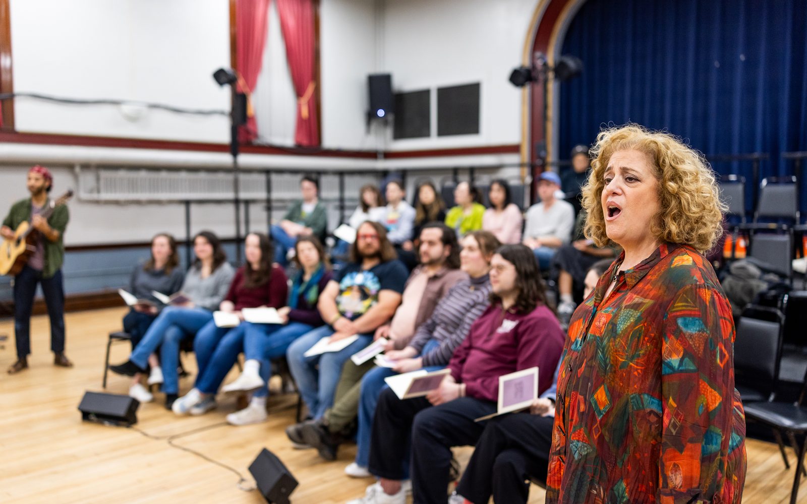 Mary Testa with curly strawberry blonde hair and in a red patterned blouse sings to one side of the audience.