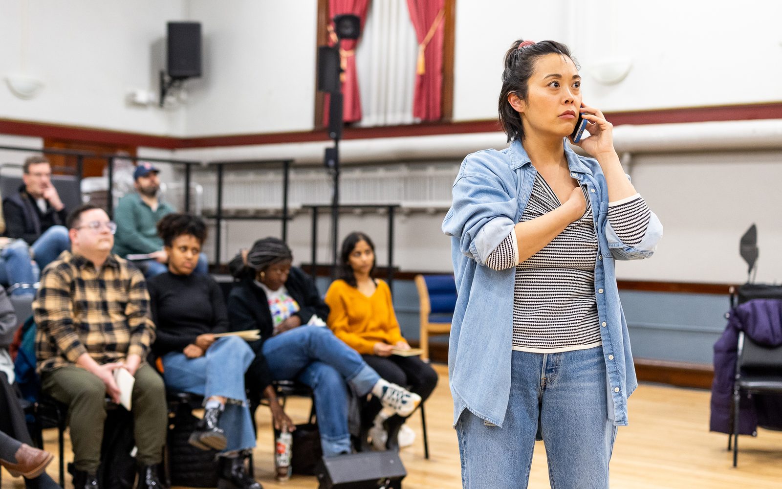 Brooke Ishibashi with her hair pulled back, wearing jeans, an open denim button-down, and a striped shirt holds a phone to her ear with one hand as her other rubs her heart.