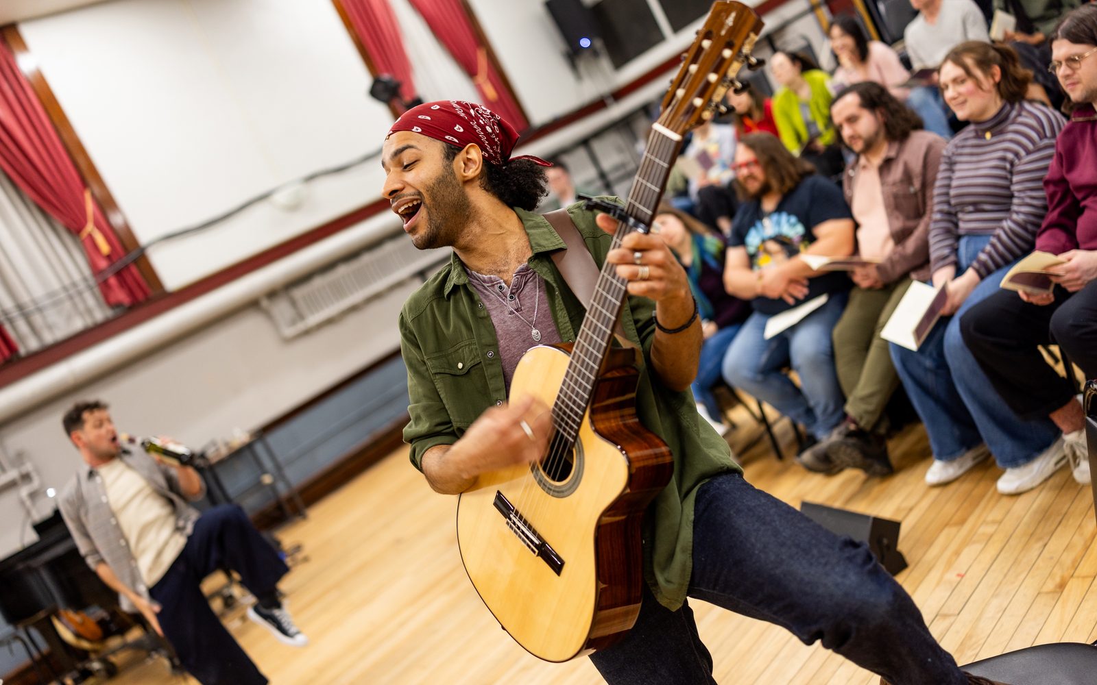 Jordan Dobson in a red bandana, green utility shirt, and grey henley sings broadly while playing a guitar.