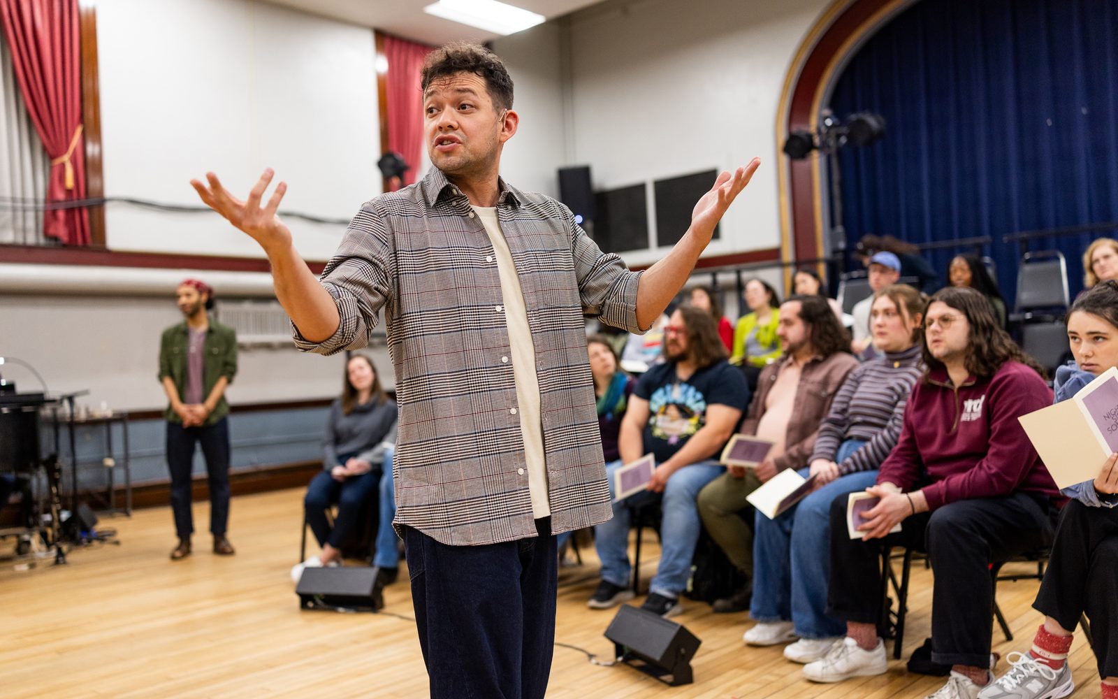 Robi Hager in an open blue and white, thin-plaid button-down raises his arm as he walks past the audience.