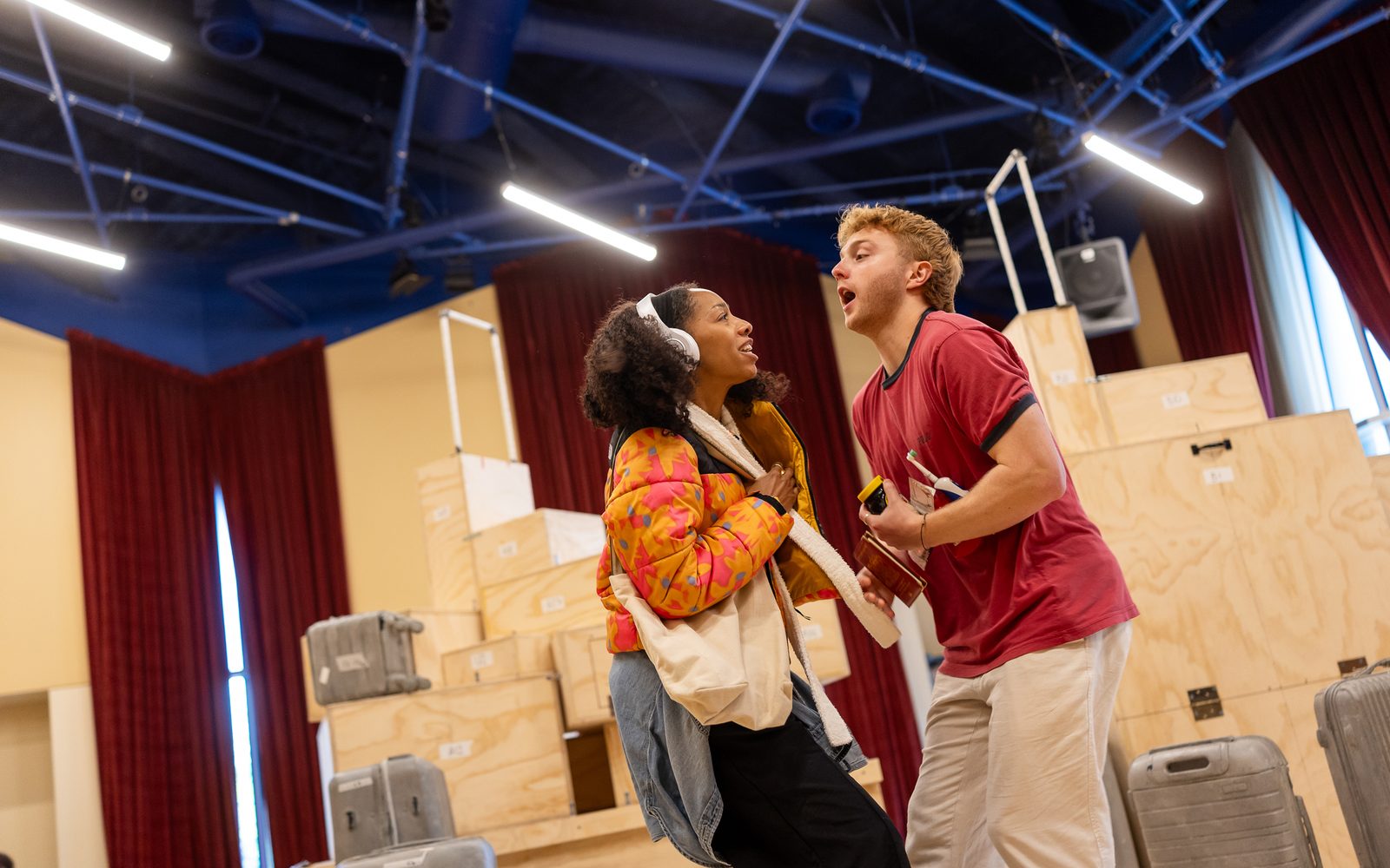 Christiani Pitts in an orange patterned puffer jacket and wearing headphones bumps into Sam Tutty in a red T-shirt and carrying several small items.