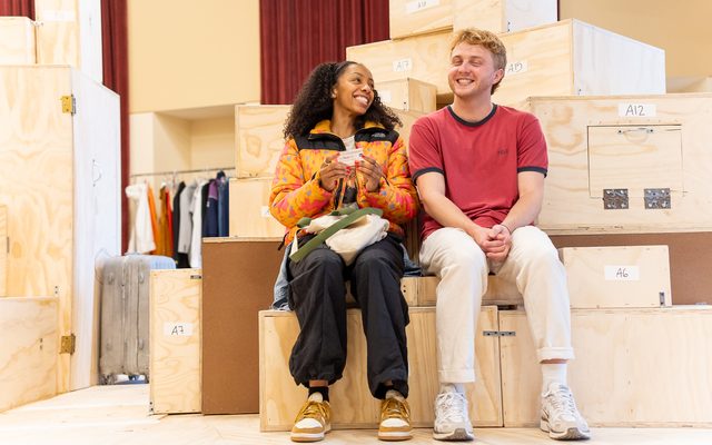 Christiani Pitts in an orange patterned puffer jacket and Sam Tutty in a red T-shirt sit on wooden blocks. He smiles towards the camera and she smiles at him.