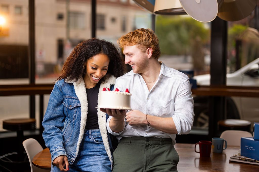 Inside a modern cafe, Christiani Pitts in a lined denim jacket and jeans looks at a white frosted cake that Sam Tutty, in a striped button-down and dark green chinos, holds while looking at her.