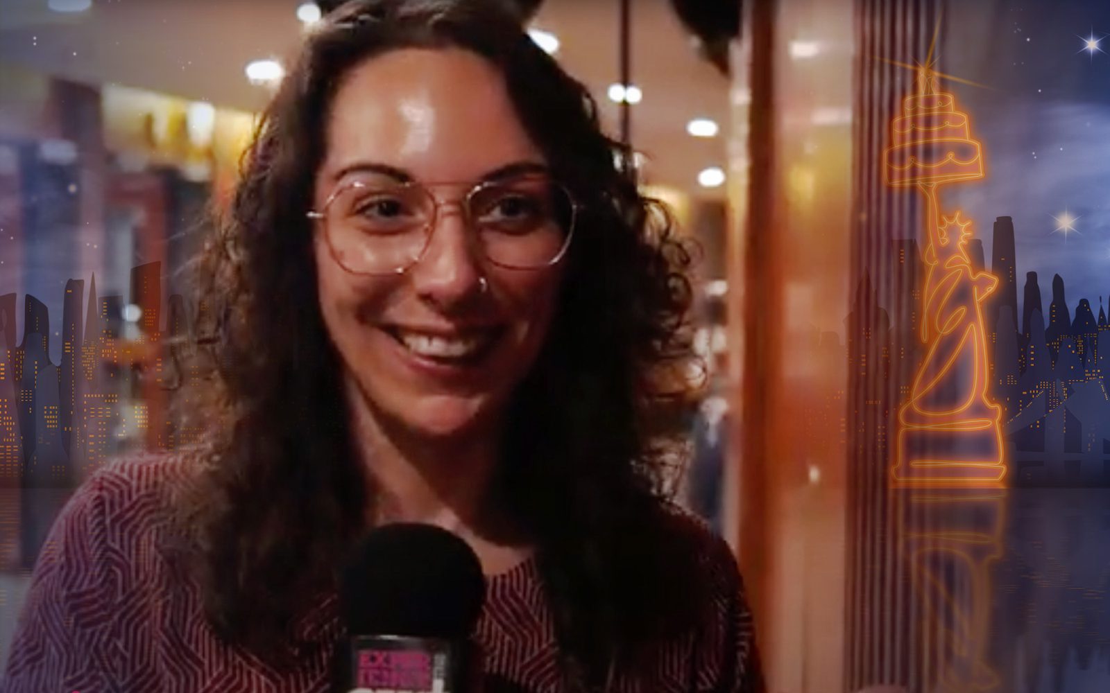 A woman with dark wavy hair and glasses speaks into a microphone.