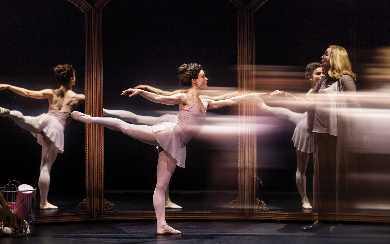 Melanie Moore in a muted pink leotard poses in first arabesque, with one leg lifted behind her. Kate Jennings Grant, standing to her right in a long beige cardigan, grasps a finger on Nina's outstretched hand. Nina's reflection can be seen in the mirrors surrounding them.