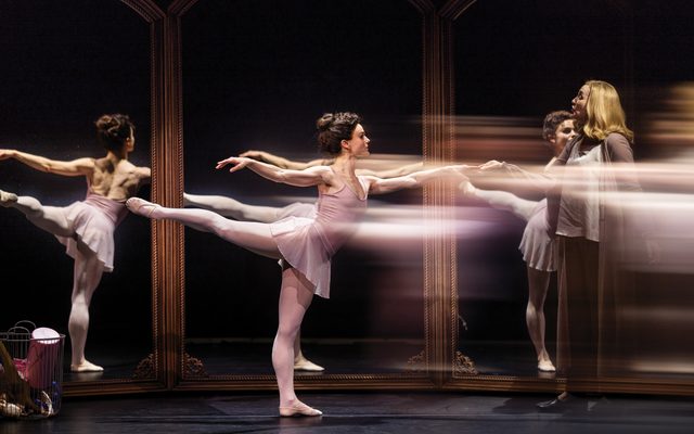 Melanie Moore in a muted pink leotard poses in first arabesque, with one leg lifted behind her. Kate Jennings Grant, standing to her right in a long beige cardigan, grasps a finger on Nina's outstretched hand. Nina's reflection can be seen in the mirrors surrounding them.