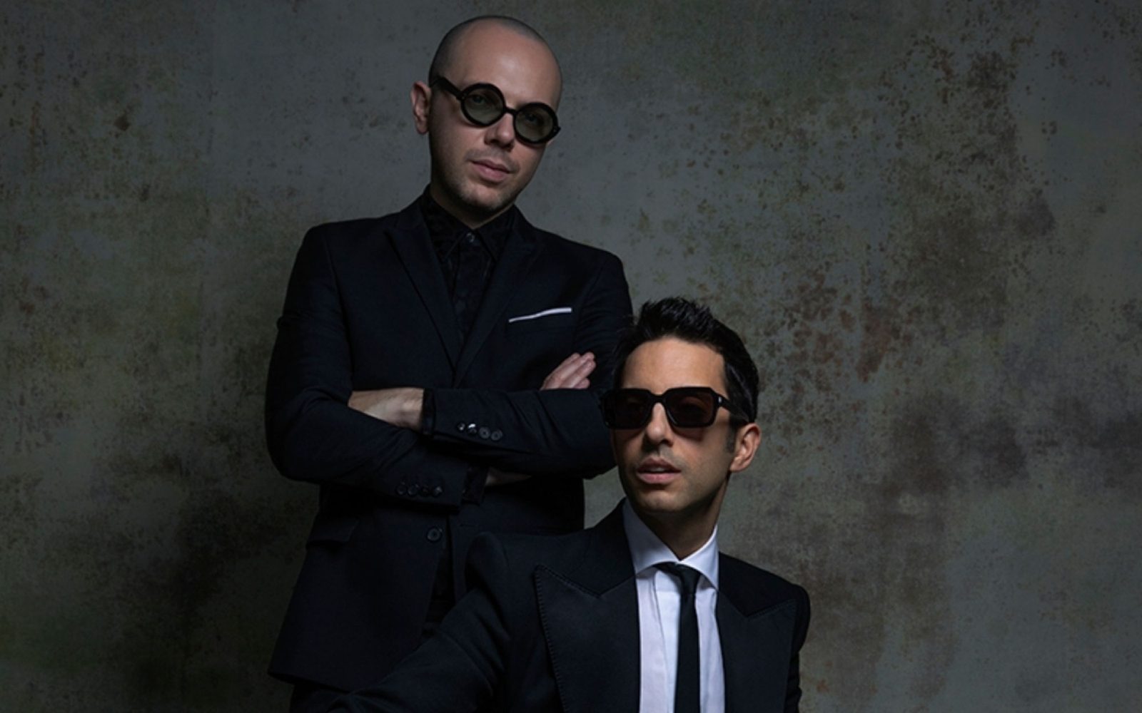 Ian Axel in a black suit and dark glasses stands with arms crossed behind Chad King's seated in a suit and dark glasses against a grey backdrop.