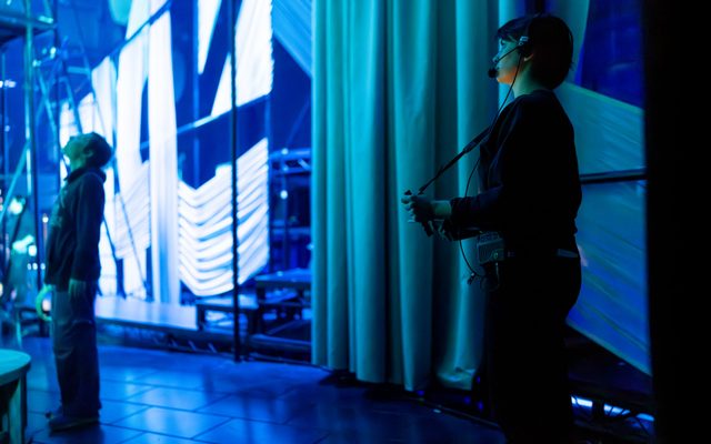 Two stage hands are backstage in bathed in blue shadow, in front a brightly lit wall of fabric panels.