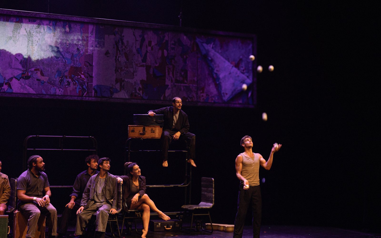 An acrobat stands downstage juggling seven balls, while other performers sit behind him on set pieces arranged like a train car.