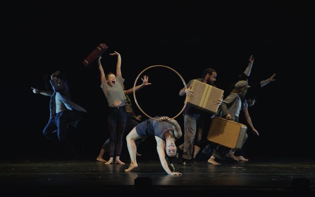 An acrobat performs a backbend and holds a hula hoop above her, as other performers break away to either side of her carrying luggage.