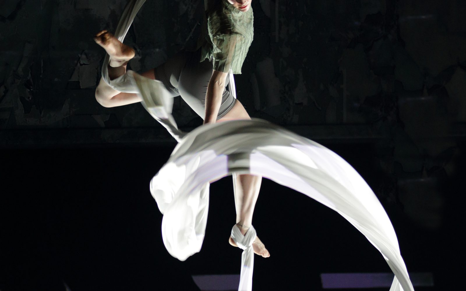 Against a dark backdrop, an acrobat suspended in midair by white silks swirls one in front of her.