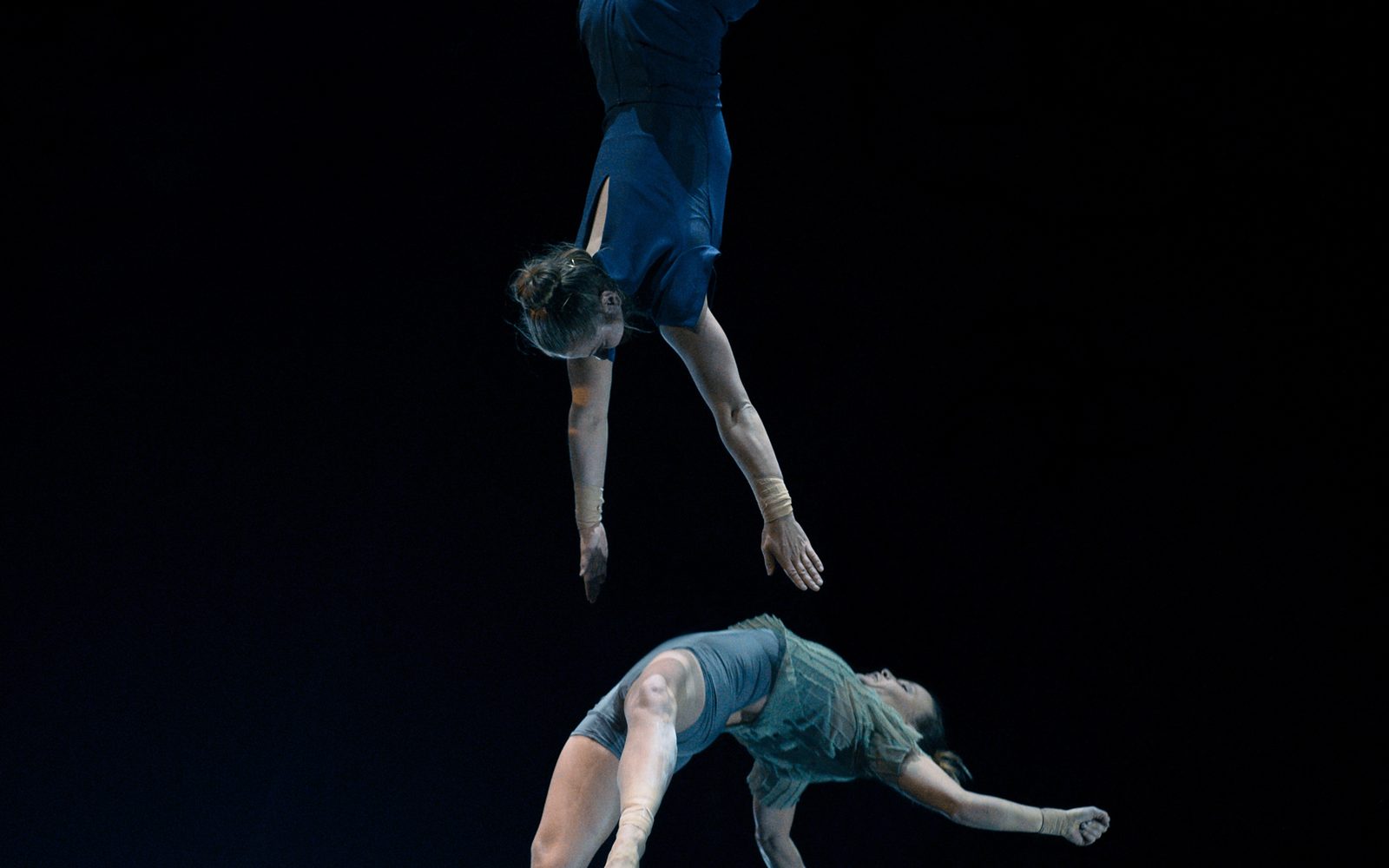 An acrobat in blue hangs down by the legs from a trapeze catching another acrobat thrown upward by a group below.
