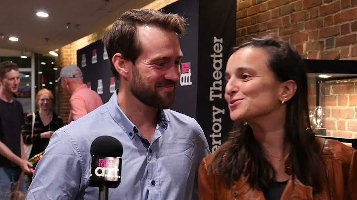 A bearded brunet man smiles at a brunette woman speaking into a microphone in the A.R.T. lobby.