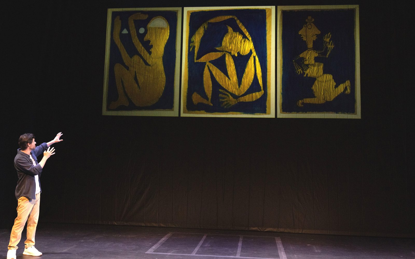 Sam Kissajukian stands downstage, turning to gesture at a projection of his painting on a screen behind him. It is a triptych of textured yellow-gold cutouts on a navy background, reminiscent of Matisse.