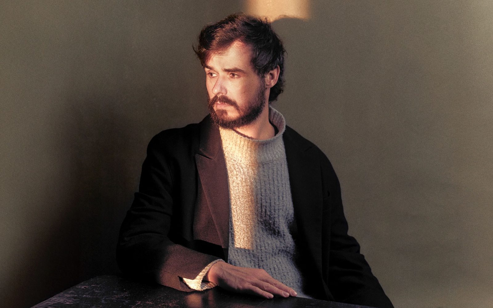 Sam Kissajukian with brown hair and a bushy beard and mustache, sits in a grey cable-knit sweater and black wool coat at a table, and glances to the left into a sliver of light.