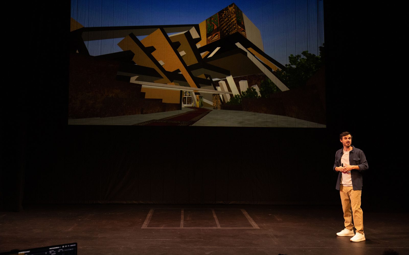 Sam Kissajukian stands downstage with a painting of his projected on a screen behind him. The paintings is a quasi-Brutalist fantastical structure in brown, tan, black, and grey against a blue sky.