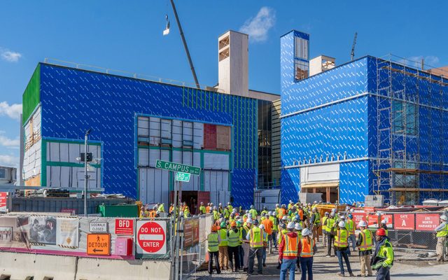 A crowd of people in hardhats and reflective vests gathers outside of the under-construction Goel Center, with two large walls in blue housewrap, to watch a crane lift a piece of timber above the building.