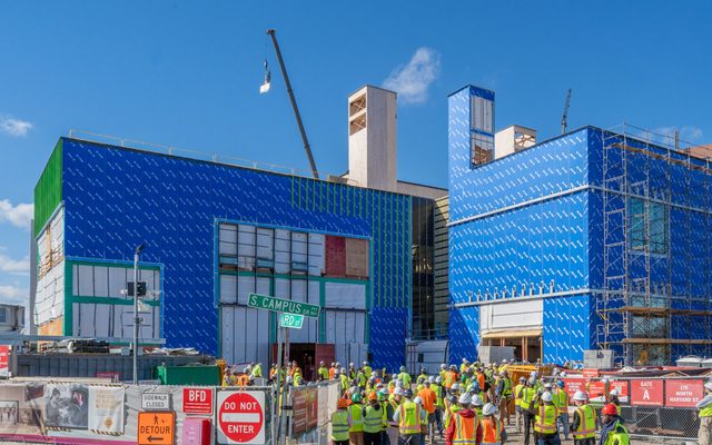 A crowd of people in hardhats and reflective vests gathers outside of the under-construction Goel Center, with two large walls in blue housewrap, to watch a crane lift a piece of timber above the building.