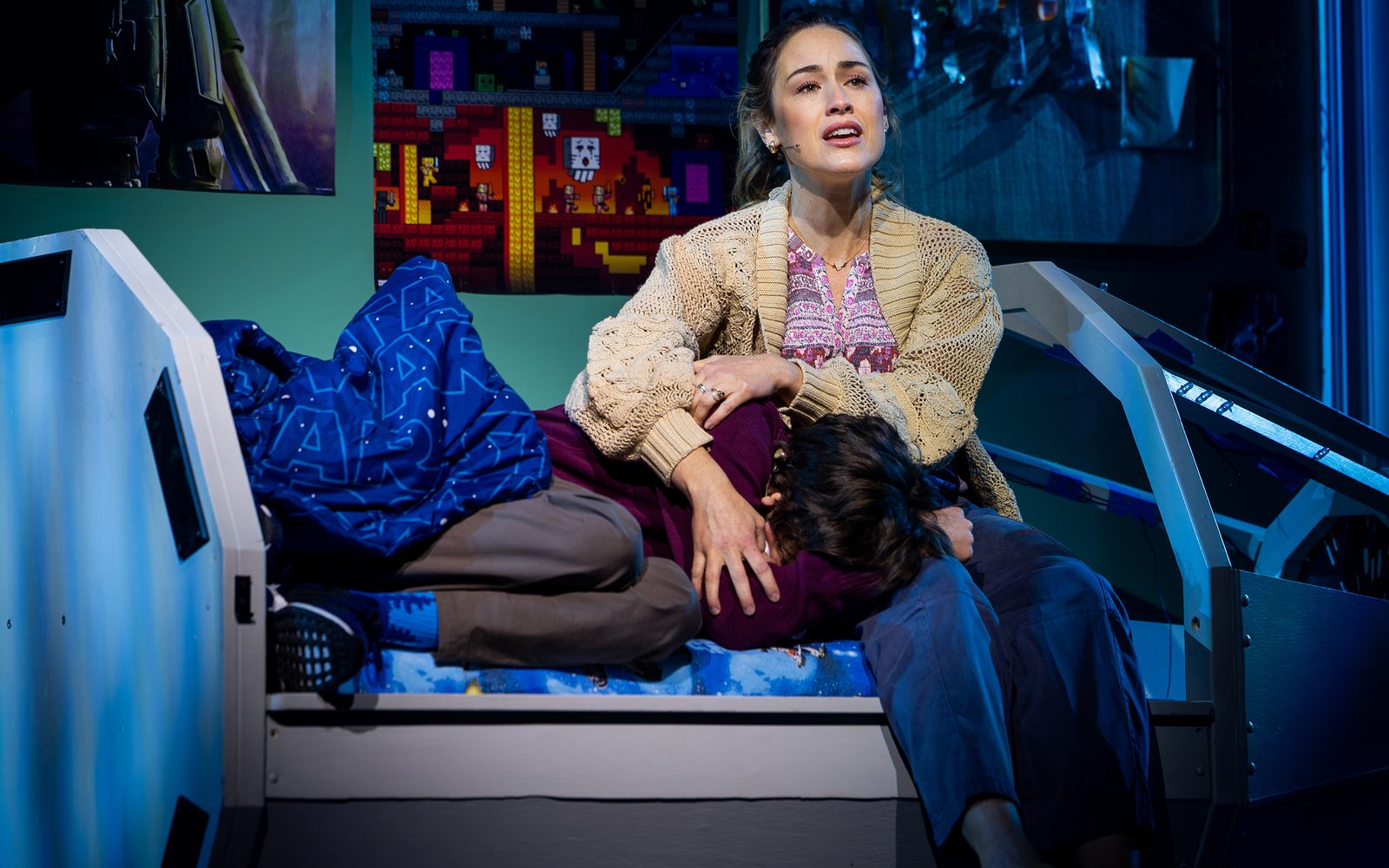 Alison Luff in a beige knit cardigan sits on the edge of a bed holding Garrett McNally, who is curled up next to her.