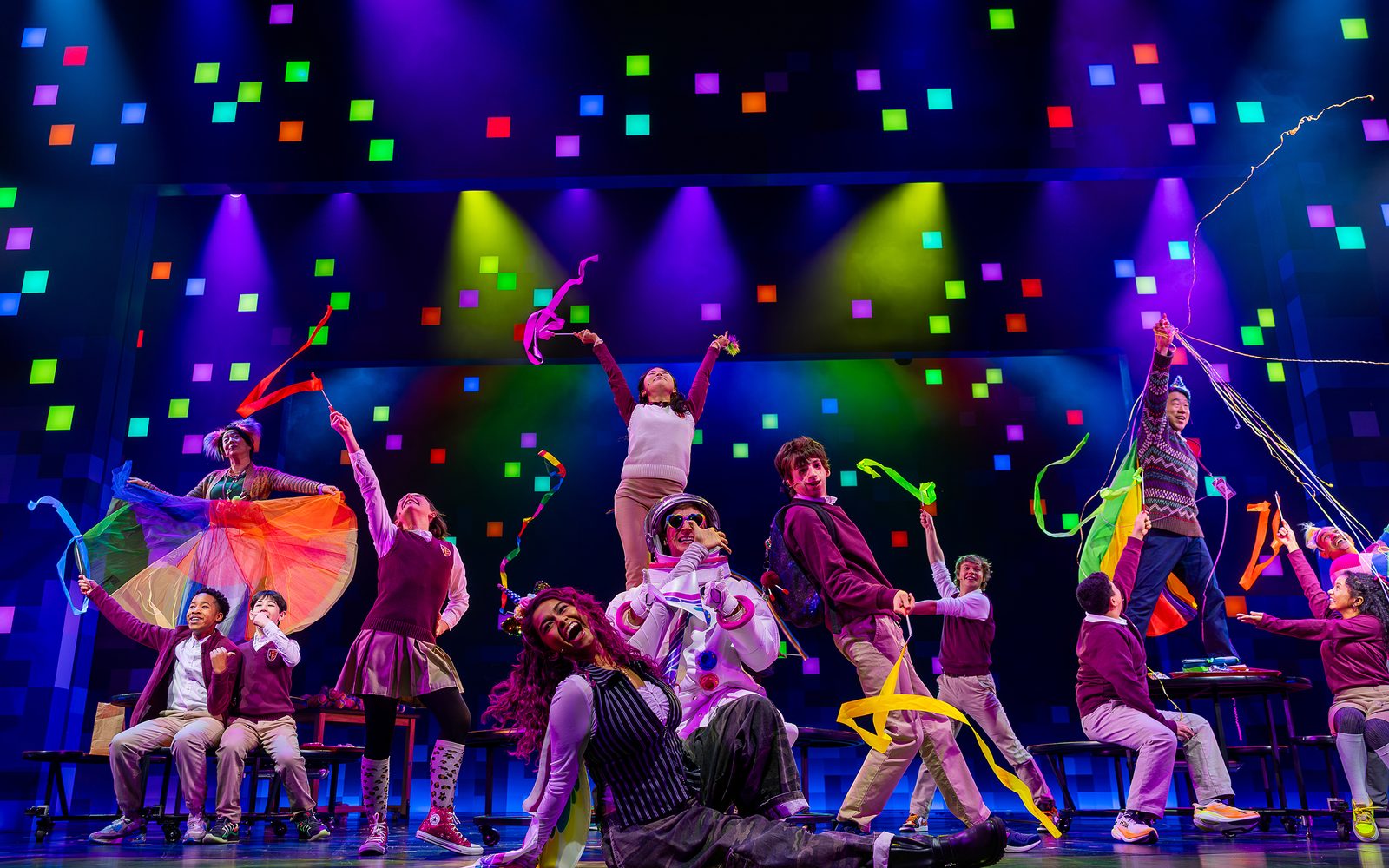 Members of the cast of Wonder pose with colorful streamers against a background with multicolored lights.