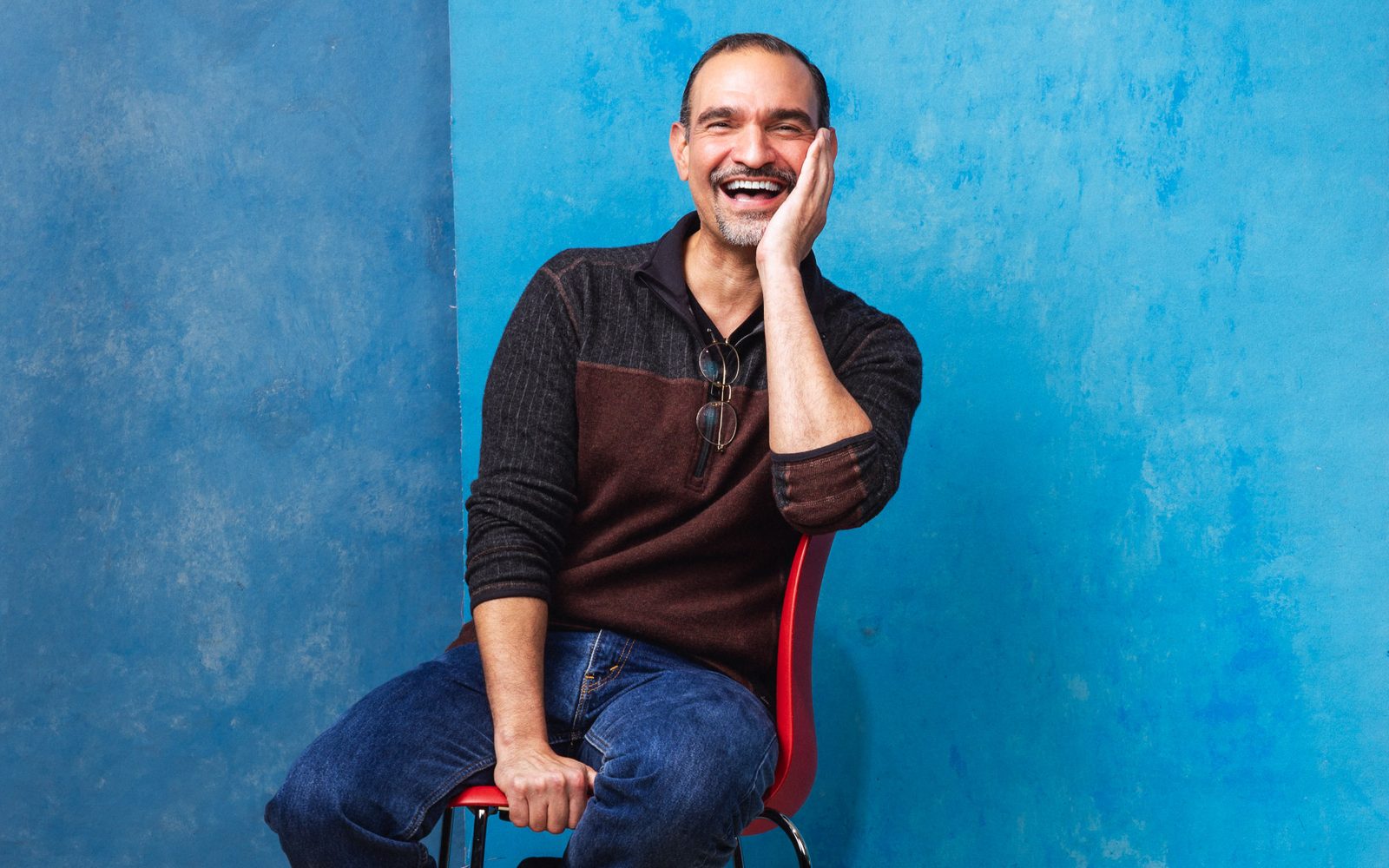 Javier Muñoz sits in a chair sideways against a blue background, smiling with his arm supporting his head.