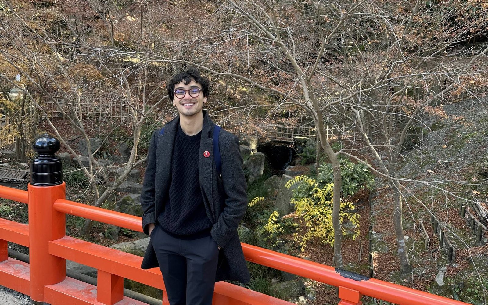 Diego Cordova in black pants, shirt, and jacket, smiles on a red Japanese bridge.
