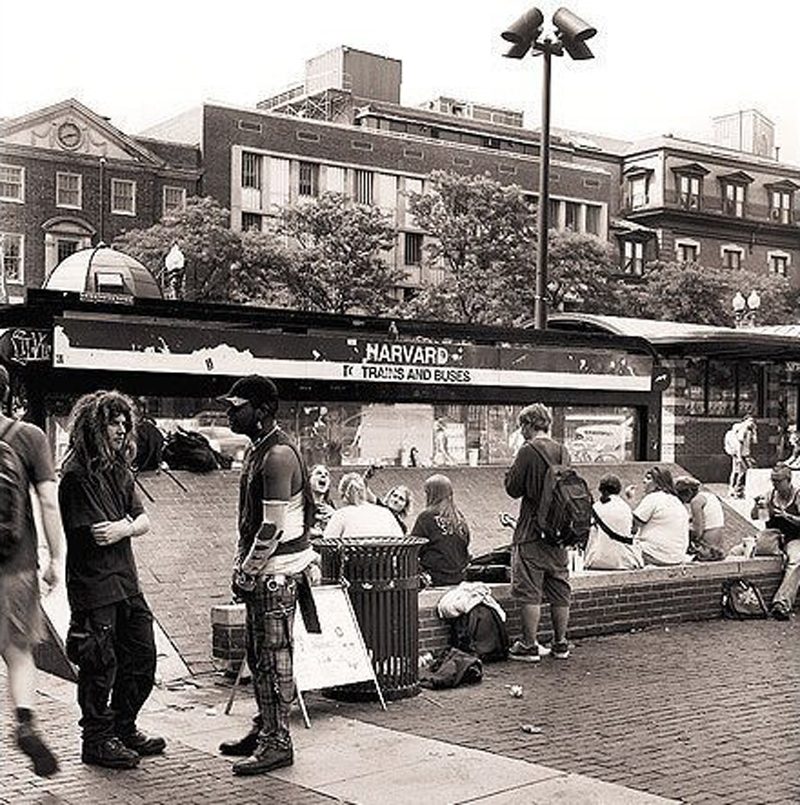 A black-and-white photo of a variety of young people sitting and standing around the sunken brick area of behind the Harvard subway head house.