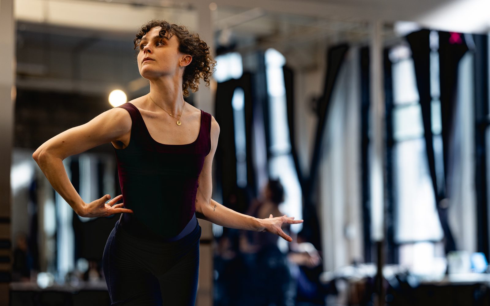 Melanie Moore looks left with one hand on her hip, and the other raised at her side in a brightly lit room.