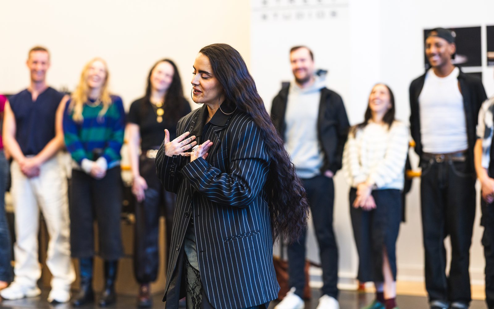 Sonya Tayeh, with her long hair down and wearing a navy pinstripe jacket, stands in front of a group of people speaking and gesturing at her chest.