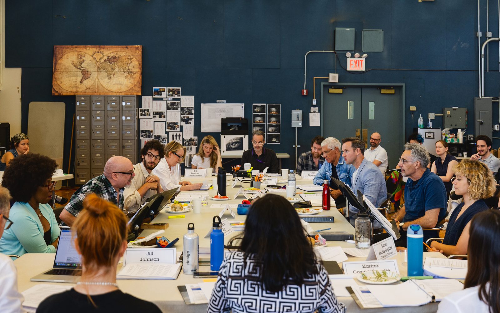 A view of the whole company sitting around a large table. Strewn about are scripts, laptops, water bottles, and plates of food, amongst other things.
