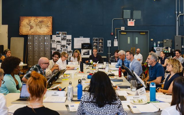 A view of the whole company sitting around a large table. Strewn about are scripts, laptops, water bottles, and plates of food, amongst other things.
