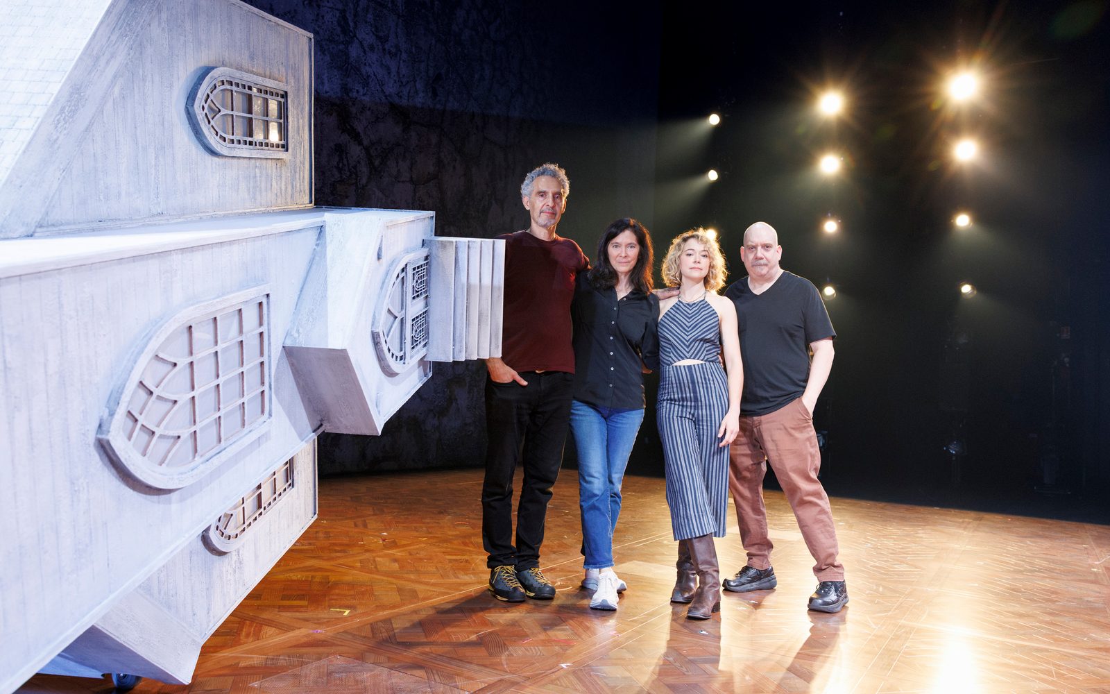 John Turturro, Diane Paulus, Tatiana Maslany, and Paul Giamatti pose together in a line onstage next to a playhouse-sized church tipped on its side.