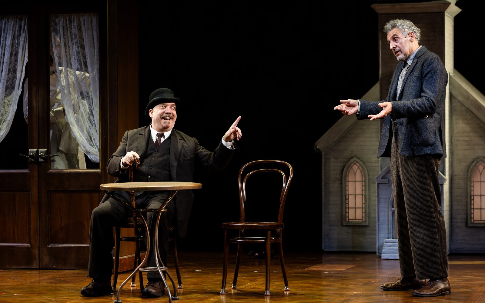 Paul Giamatti in a three-piece suit and bowler hat sits at a cafe table smiling and pointing at John Turturro, who stands shrugging in a wool blazer.