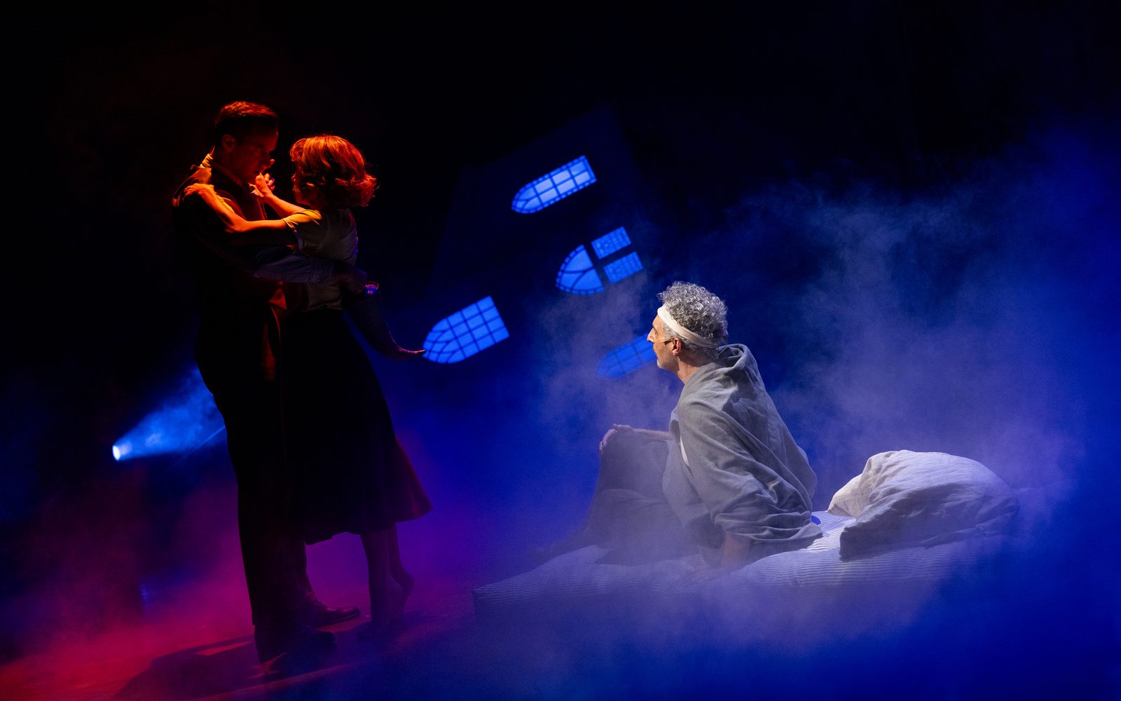 John Turturro with a bandage wrapped around his head rises from a cot surrounded by fog, staring at Alex Ross and Tatiana Maslany embracing in the shadow of a red light.