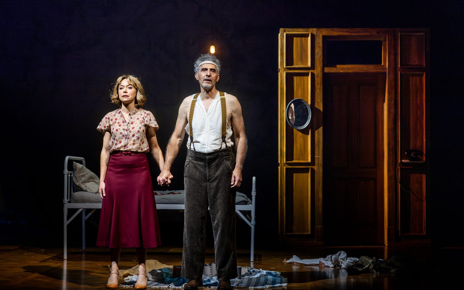 Tatiana Maslany in a muted floral blouse and John Turturro in an undershirt and suspenders stand in front of a cot, clasping hands and staring out expressionlessly.