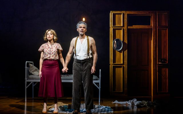 Tatiana Maslany in a muted floral blouse and John Turturro in an undershirt and suspenders stand in front of a cot, clasping hands and staring out expressionlessly.