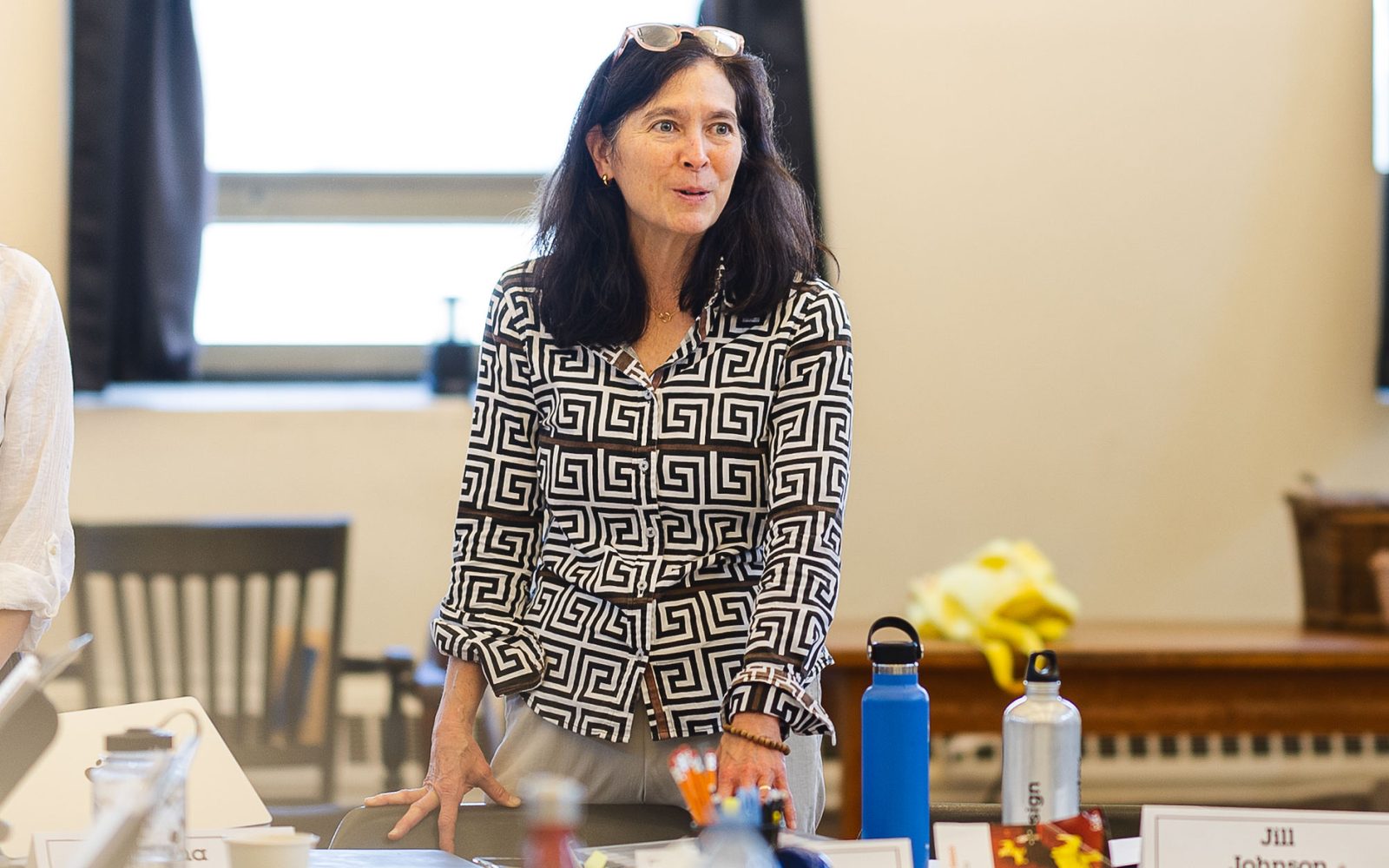 Diane Paulus with dark brown hair, a black, white, and red geometric patterned blazer, and white glasses atop her head, stands to address the table.