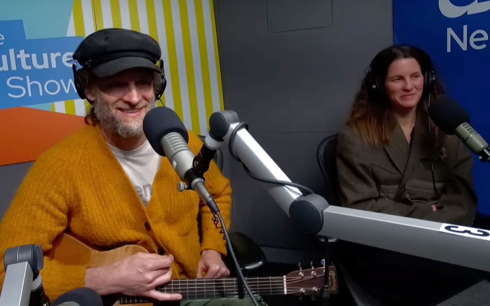 Chadiwkc Stokes in a gray fiddler cap and orange cardigan sitting in front of a studio mic, with Sybil Gallagher in a brown jacket sitting in front of a mic to the right.